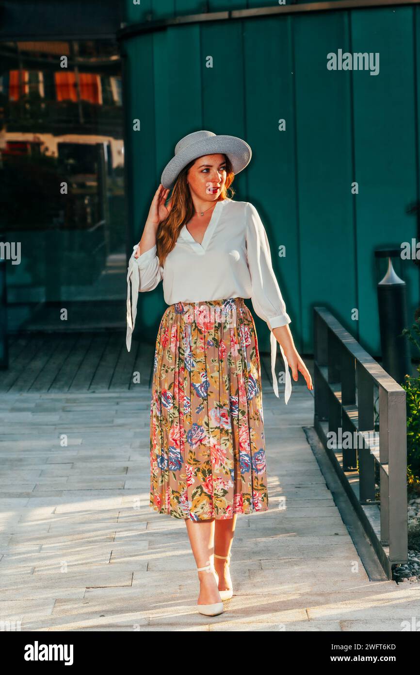 Giovane donna alla moda con cappello, lunghi capelli biondi, blusa bianca e fiorita, lunga gonna a pieghe. Foto Stock