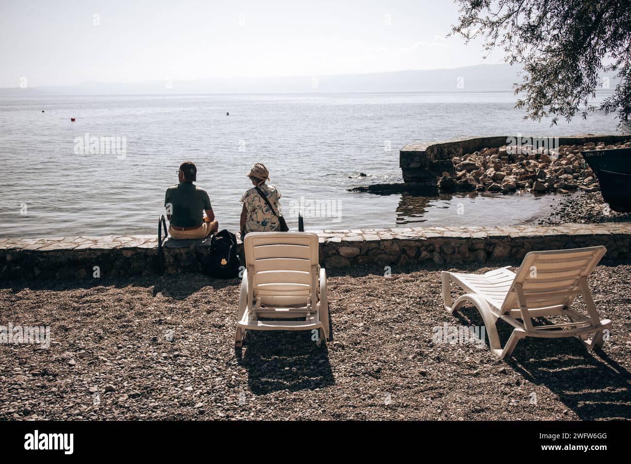 Due persone sedevano accanto all'acqua sulle rive del lago di Ocrida nella Macedonia del Nord in una bella giornata di sole, con due lettini accanto a loro sulle rocce. Foto Stock