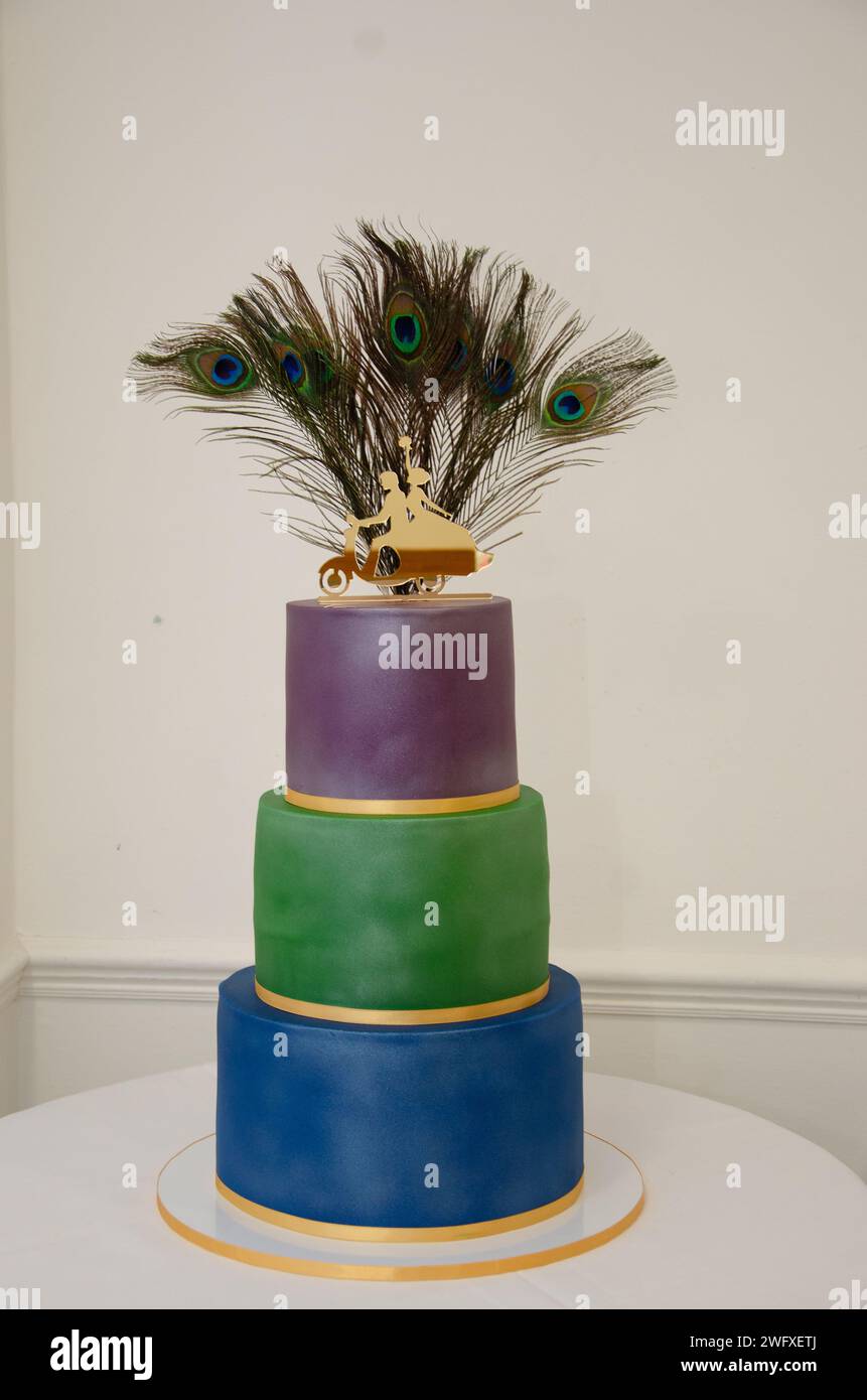 Torta nuziale con vere piume di pavone in posizione verticale sullo strato superiore di una torta a 3 livelli con ogni strato di colore viola blu e verde. La torta ha la glassa d'oro. Foto Stock