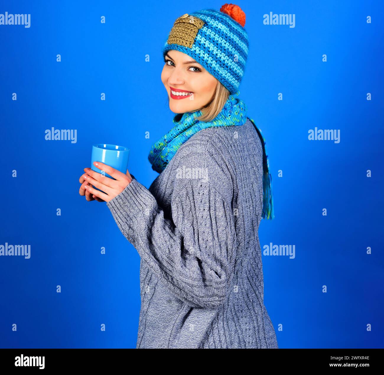 Bella donna elegante in abiti alla moda con una tazza che si gusta una bevanda calda nella caffetteria. Ragazza sorridente con maglione, cappello e sciarpa che beve caffè caldo o. Foto Stock