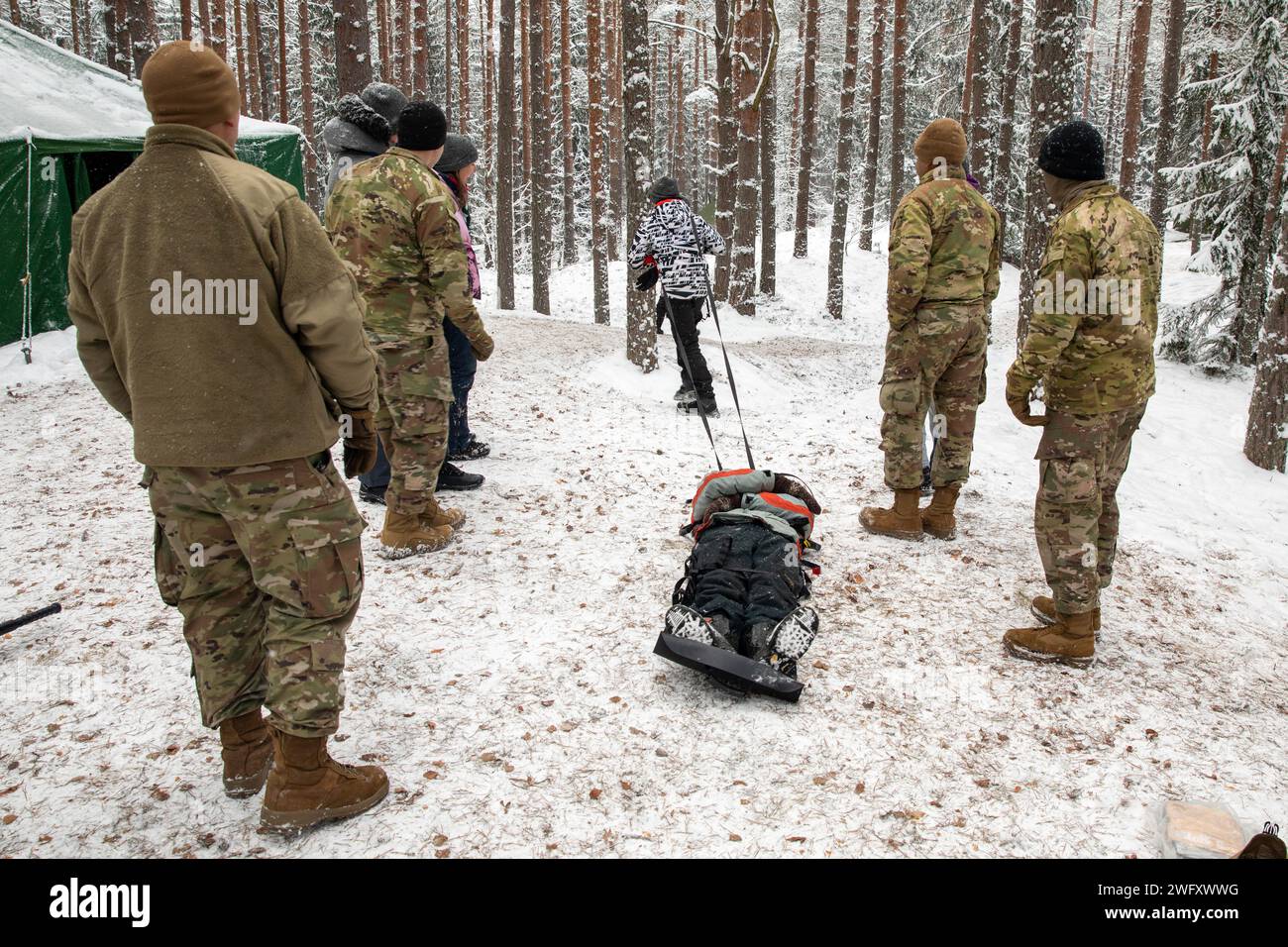 I medici dell'esercito degli Stati Uniti della 3rd Infantry Division Artillery and Civil Affairs Soldiers del 432nd Civil Affairs Battalion a sostegno della 3rd Infantry Division guardano da bambino che frequenta un campo invernale per bambini lettoni usa una lettiera per trasportare un paziente simulato vicino a Jelgava, Lettonia, 6 gennaio. 2024. I medici hanno insegnato lezioni sulla prevenzione delle lesioni da freddo e l'uso di una lettiera. La missione della 3rd Infantry Division in Europa è quella di impegnarsi in formazione ed esercitazioni multinazionali in tutto il continente, lavorando al fianco degli Alleati della NATO e dei partner di sicurezza regionali per fornire forze di combattimento credibili Foto Stock