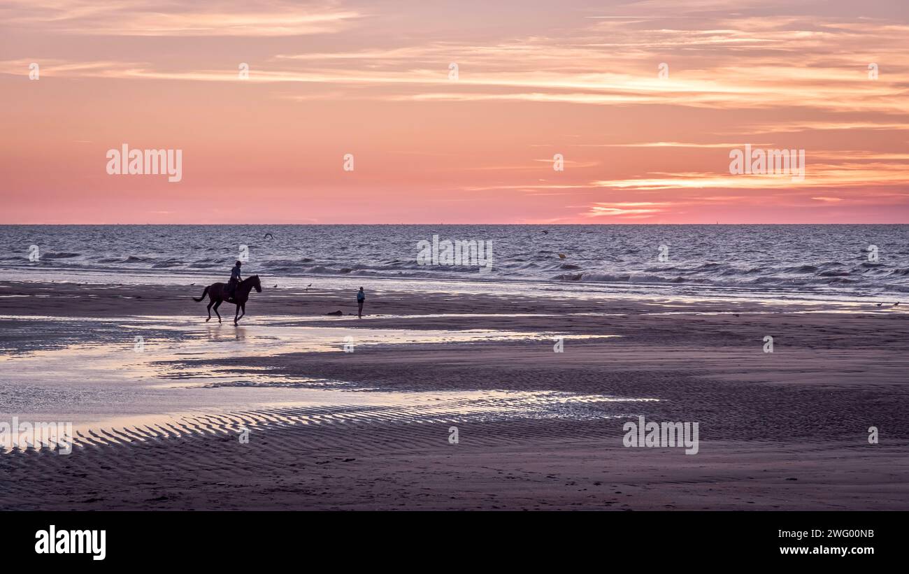 Due persone a cavallo lungo la spiaggia sabbiosa durante un pittoresco tramonto Foto Stock