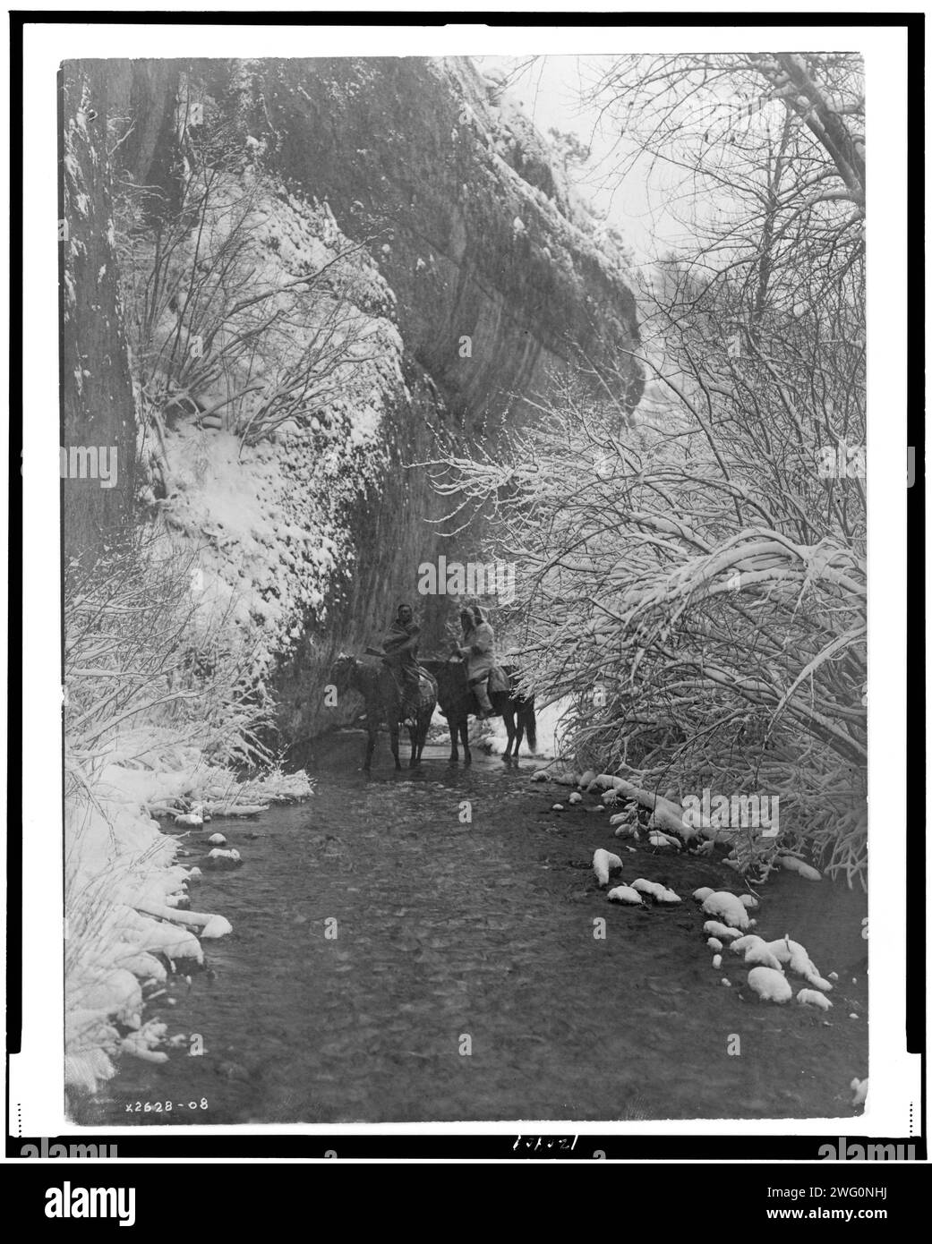 Si avvicina all'inverno-Apsaroke, c1908. Due Crow Indiani a cavallo in un ruscello poco profondo fiancheggiato da alberi innevati, sotto la scogliera rocciosa. Foto Stock