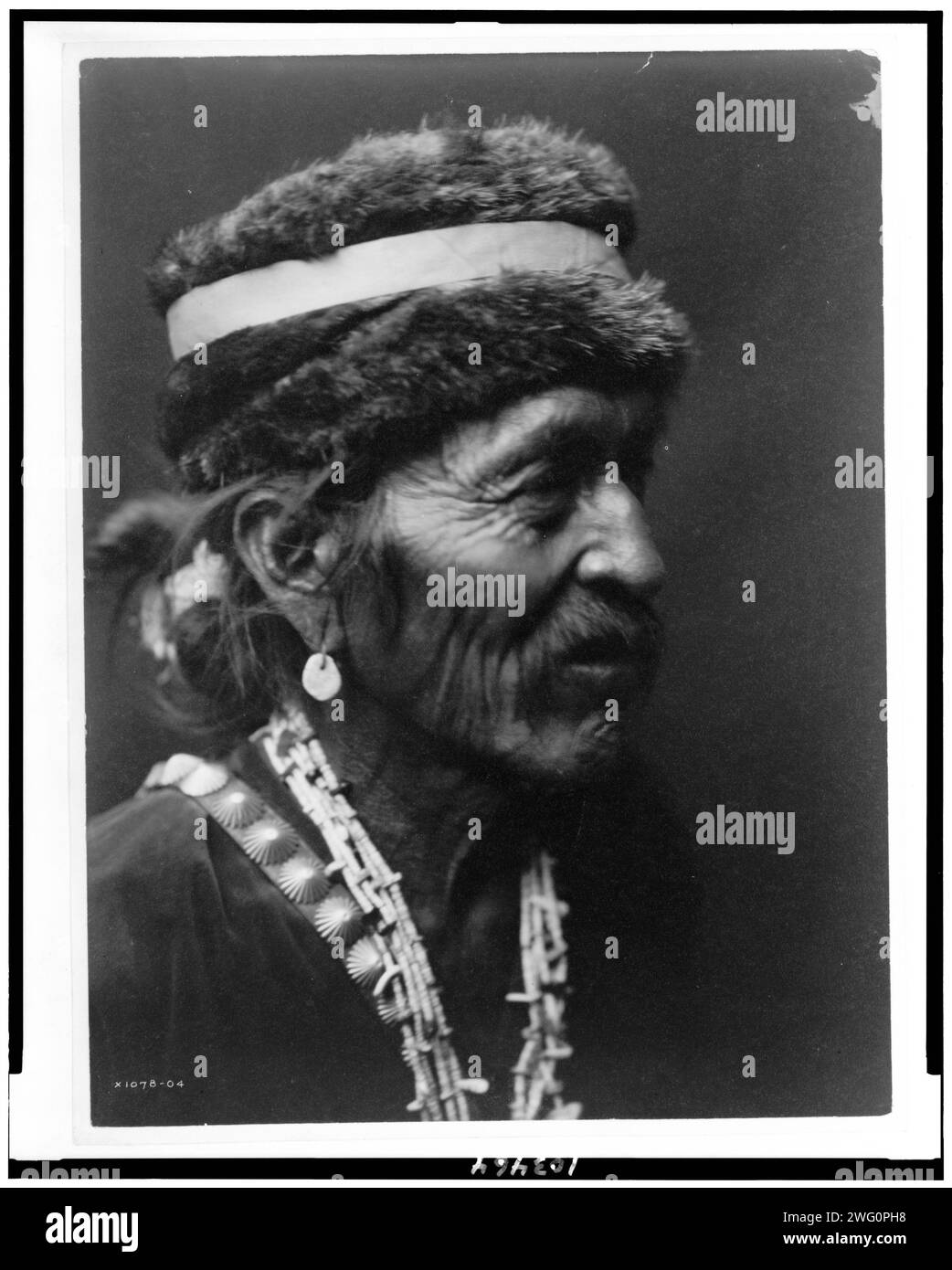 Navajo con berretto in pelliccia, c1905. Ritratto dell'uomo Navajo con la testa e le spalle rivolto verso destra, con berretto in pelliccia, orecchino turchese, collana con conchiglie, tracolla con bottoni in argento e camicia in velluto. Foto Stock