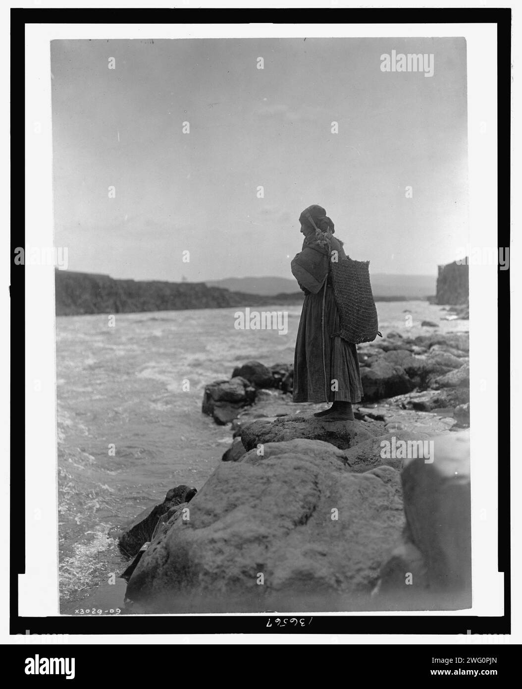 Il portapesci-Wisham (cioè Wishram), c1910. Un indiano Tlakuit in piedi sulla roccia ai bordi delle acque, indossa un abito lungo la caviglia e una borsa grande sulla schiena, con cinturino appoggiato alla corona della testa. Foto Stock