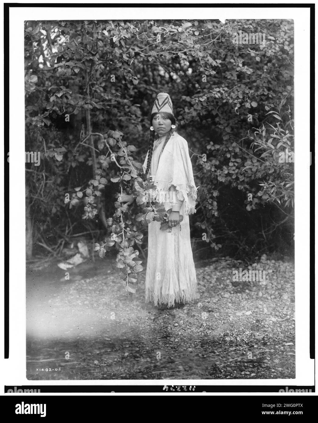Una bella fanciulla, c1905. Nespelim Woman si trova sul bordo dell'acqua, alberi dietro di lei. Foto Stock