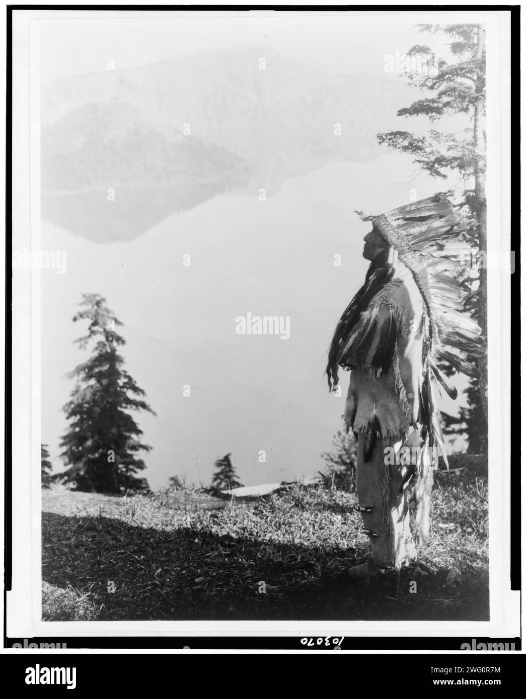 Pregare gli spiriti al Crater Lake-Klamath, c1923. Capo indiano di Klamath in piuma che si trova sulla montagna che si affaccia sul lago Crater, Oregon. Foto Stock