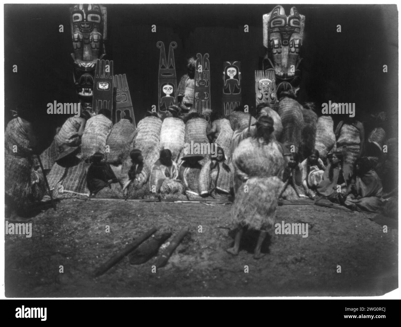 Un incidente della Winter Dance-Nakoaktok, c1914. Gente di Kwakiutl, alcuni inchinandosi prima dei pali totemici sullo sfondo, altri seduti di fronte come parte della cerimonia dei nunhlim, i quattro giorni prima della Danza invernale. Foto Stock