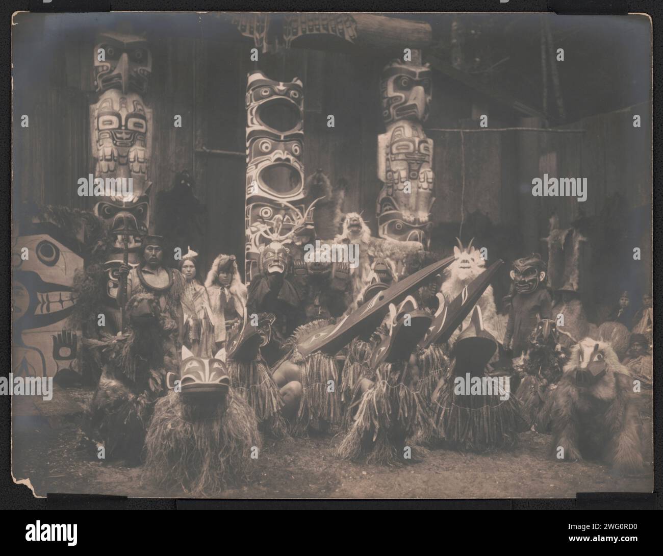 Ballerini mascherati-Qagyuhl, c1914. Durante la cerimonia invernale, i ballerini Kwakiutl indossano maschere e costumi, si accovacciano in primo piano con altri dietro di loro. Il capo dell'estrema sinistra ha lo staff di un oratore. Tre pali totem sullo sfondo. Foto Stock
