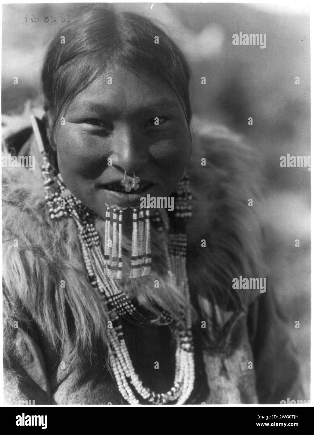 Kenowun-Nunivak, c1929. Kenowun, Nunivak Woman, ritratto testa e spalle, davanti, con parka in pelliccia, collane, orecchini, ornamenti nasali e perline sotto il labbro inferiore. Foto Stock