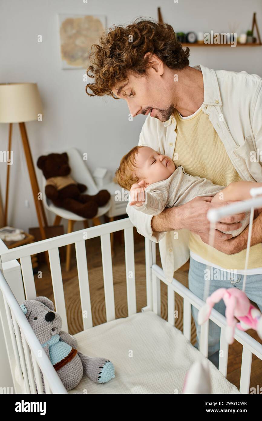 padre single riccio che tiene in braccio il suo figlio neonato vicino alla culla nella nursery, paternità e amore Foto Stock