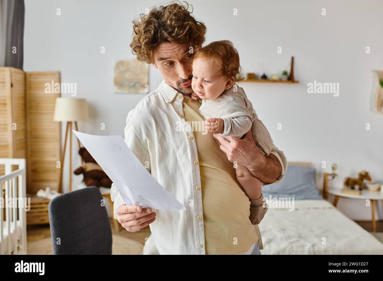 padre riccio e barbuto che tiene in braccio il bambino neonato e guarda il documento, l'equilibrio della vita lavorativa Foto Stock