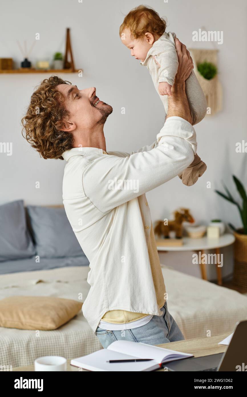 padre felice e riccio che tiene tra le braccia il figlio piccolo vicino al computer portatile e alla tazza sulla scrivania, equilibrio tra vita lavorativa Foto Stock
