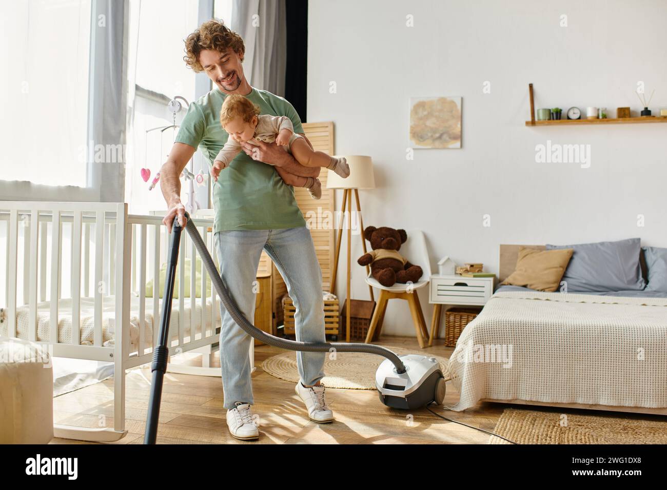 uomo multitasking lavori domestici e assistenza all'infanzia, padre che aspira pavimento in legno duro con bambino in braccio Foto Stock