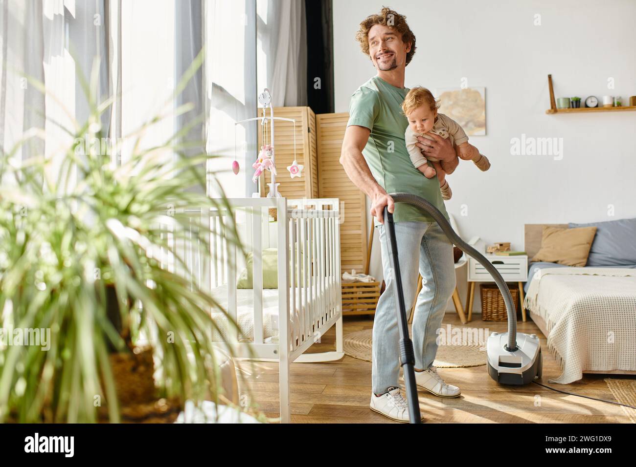 uomo multitasking lavori domestici e assistenza all'infanzia, padre single che aspira appartamento con bambino in braccio Foto Stock