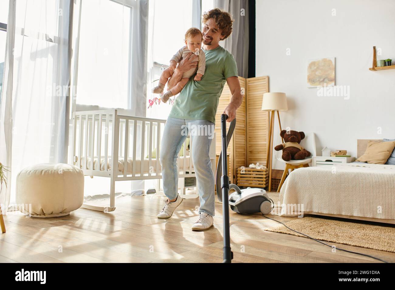 uomo felice lavoro domestico e assistenza all'infanzia in multitasking, padre che aspira il pavimento in legno duro con il bambino in braccio Foto Stock
