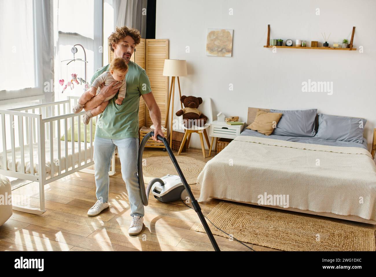 uomo che lavora in multitasking in casa e cura dei bambini, padre che aspira il pavimento in legno duro con il figlio in braccio Foto Stock