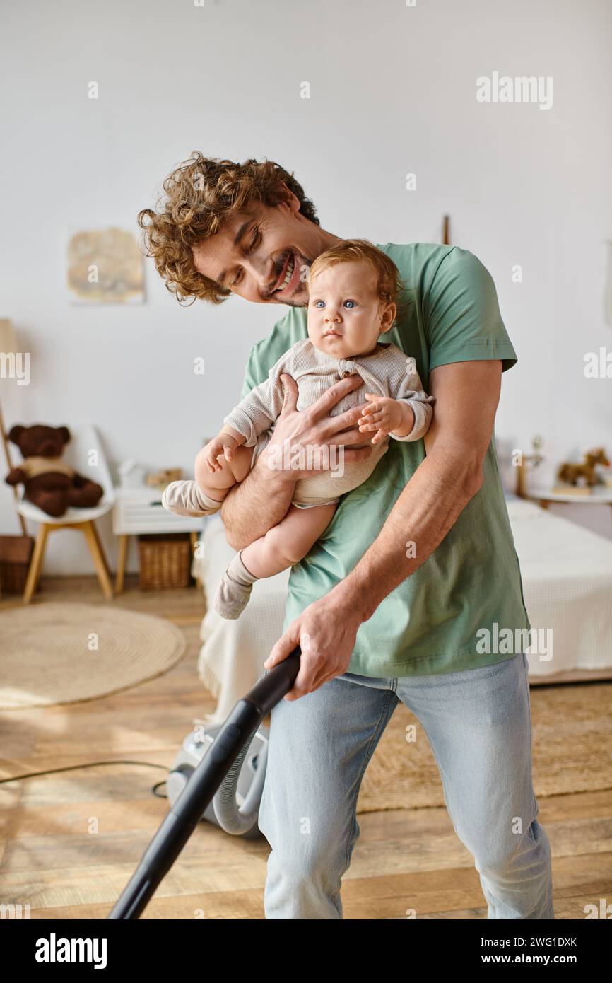 uomo che lavora in multitasking e cura dei bambini, padre felice che aspira il pavimento in legno duro con il bambino tra le braccia Foto Stock