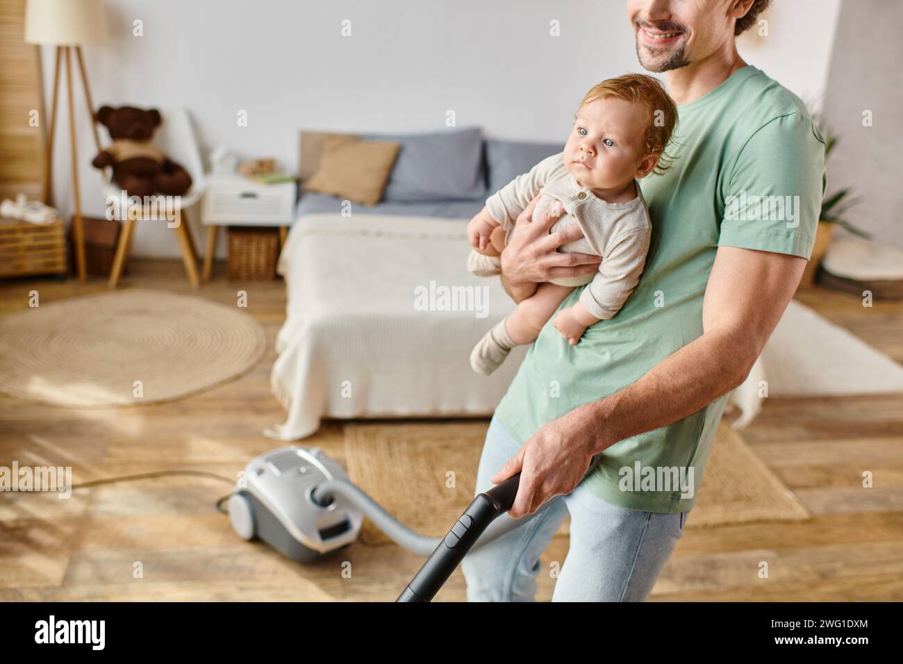 uomo corrotto lavoro domestico e assistenza all'infanzia, padre allegro che aspira casa con figlio in braccio Foto Stock