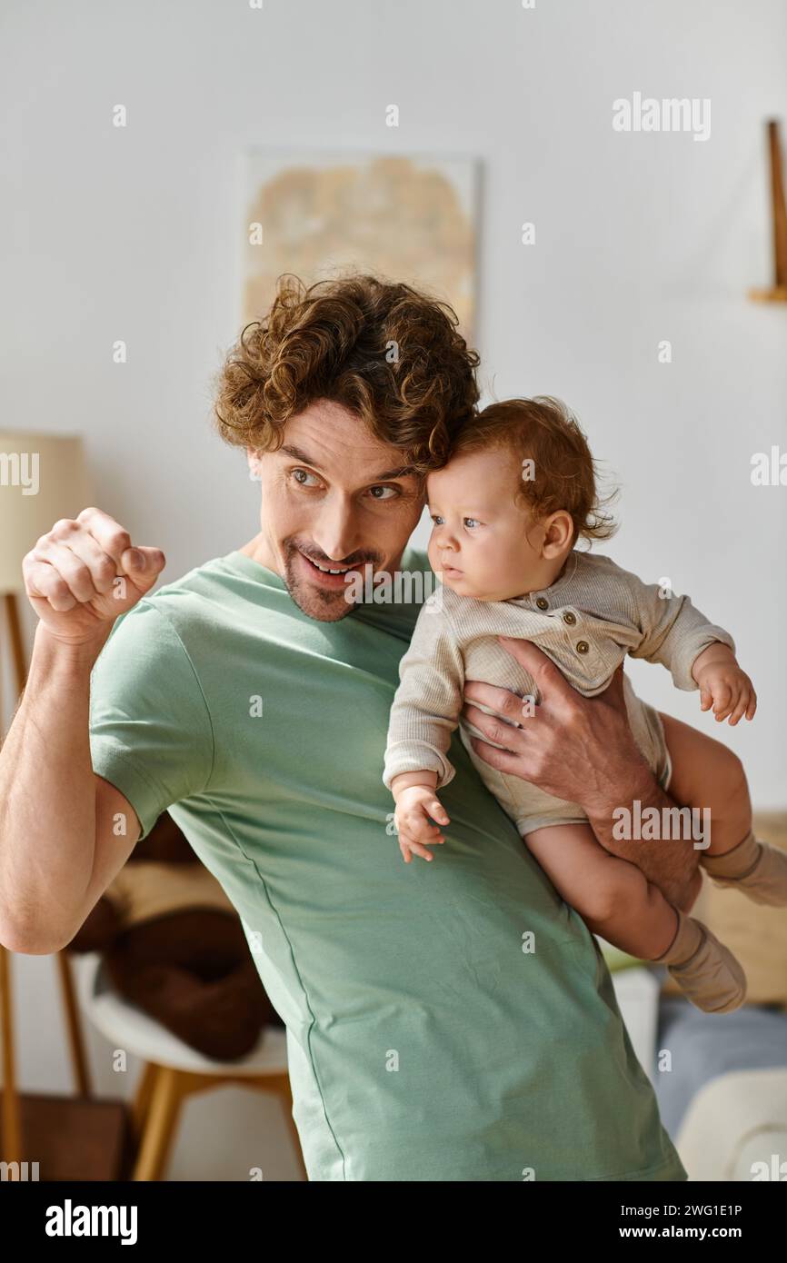 padre dai capelli ricci che tiene tra le braccia il figlio neonato e punta il dito in una camera da letto accogliente Foto Stock