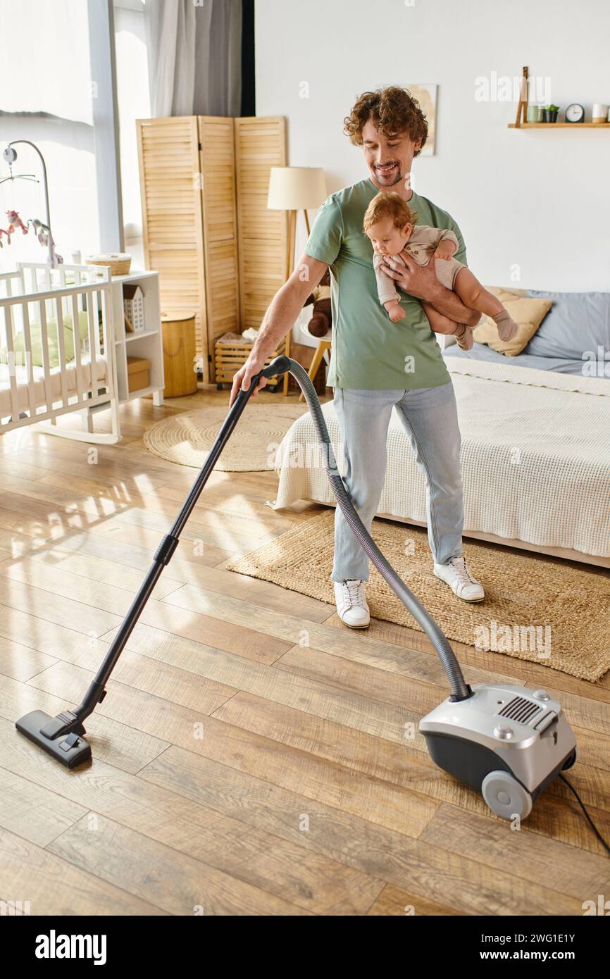 padre felice, lavoro domestico e assistenza all'infanzia, uomo che aspira la camera da letto con figlio neonato in braccio Foto Stock