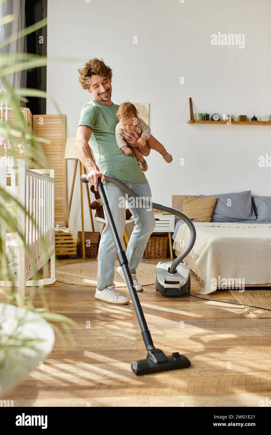uomo multitasking lavori domestici e assistenza all'infanzia, padre sorridente che aspira appartamento con bambino in braccio Foto Stock
