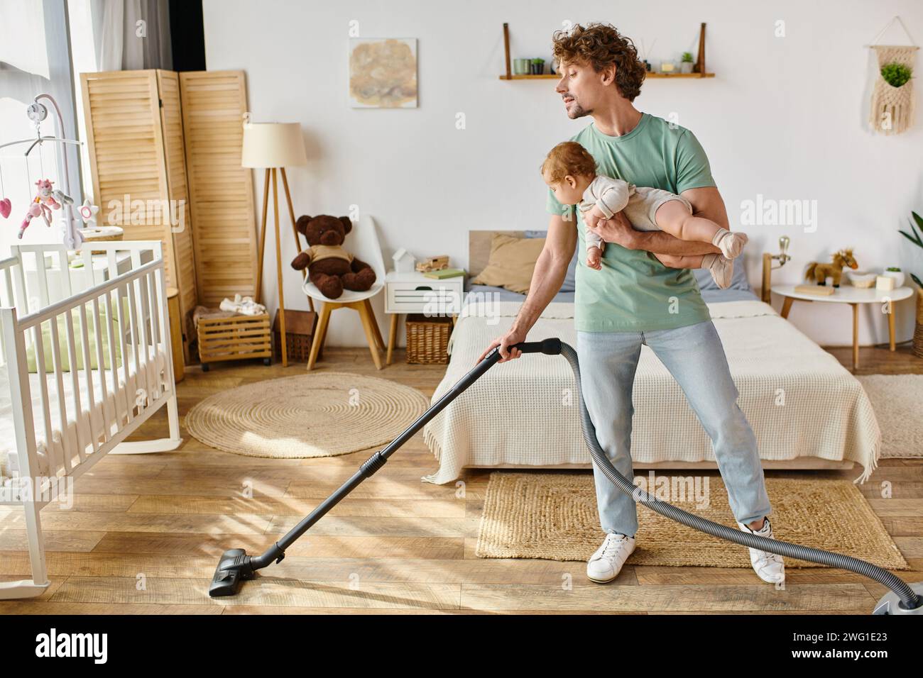 uomo riccio lavori di casa e assistenza all'infanzia, padre che aspira la camera da letto con bambino in braccio Foto Stock