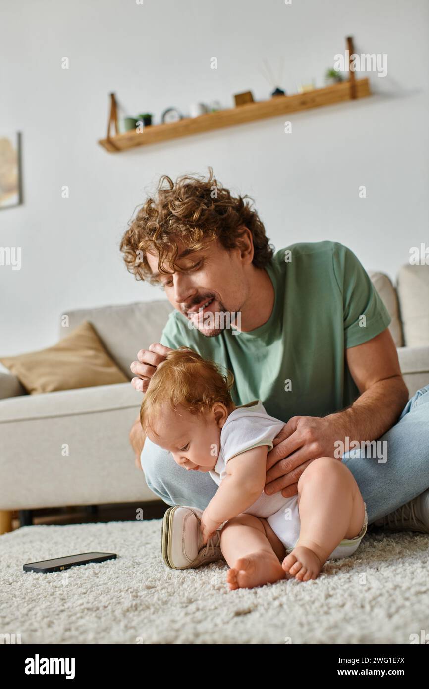 buon padre single che usa lo smartphone mentre si siede a casa con il bambino sul tappeto Foto Stock