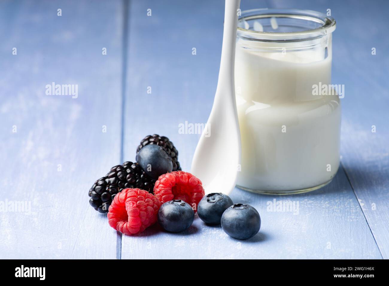 in primo piano, sul tavolo di legno, alcuni frutti di bosco freschi e un vasetto di yogurt semplice e magro con un cucchiaio di ceramica Foto Stock