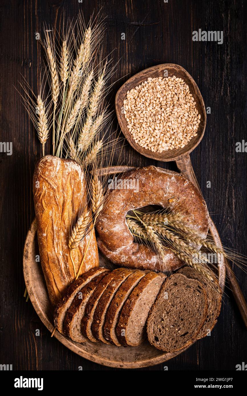 sul rustico tavolo in legno, un piatto di legno con diversi tipi di pane, con vari semi e cereali e qualche orecchie di grano. Natura morta. visualizza Foto Stock