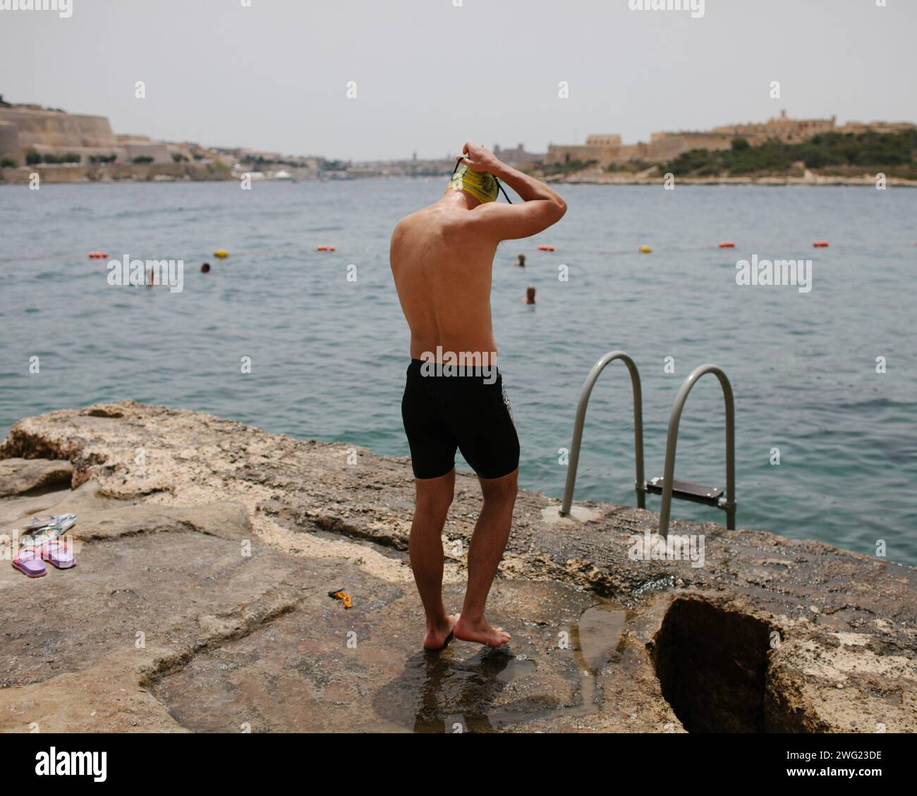 Un nuotatore si prepara ad entrare in mare alla spiaggia di Tigne Point, Malta. La Valletta può essere vista sullo sfondo. Foto Stock