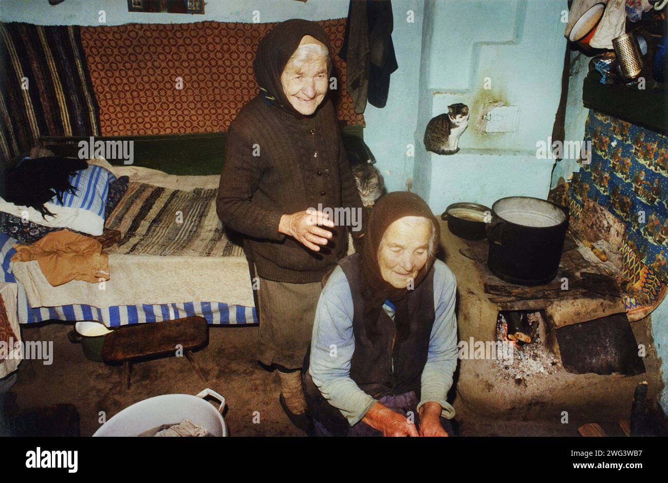 Contea di Vrancea, Romania, circa 1995. Due donne anziane in una casa rurale, con una zona cucina primitiva e un letto vicino alla fornace. Foto Stock