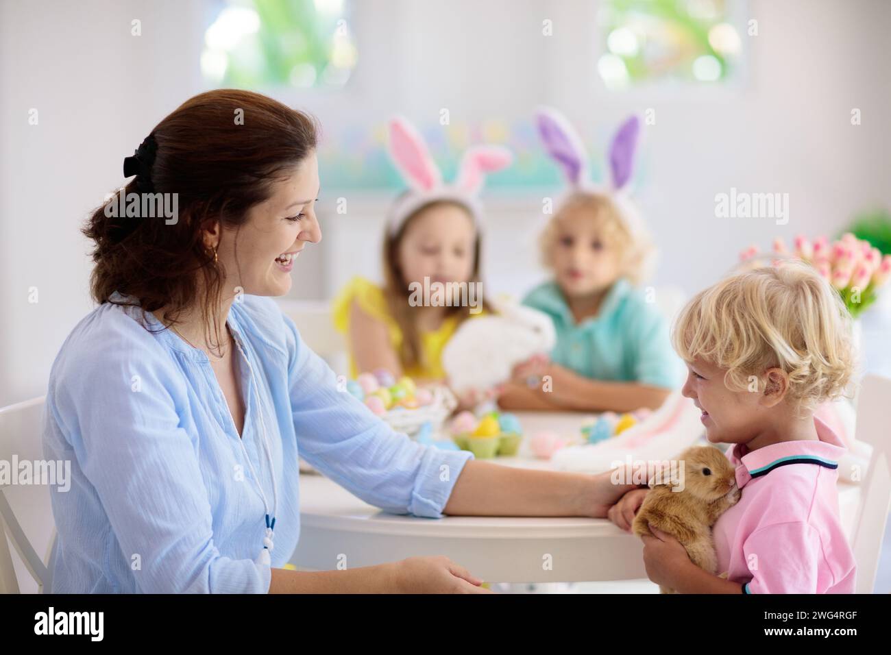 Madre e bambini con pet colore di coniglio uova di Pasqua. Mom, bambina e ragazzo con orecchie di coniglietto di tintura e pittura per uovo di Pasqua Caccia in bianco stanza soleggiata Foto Stock