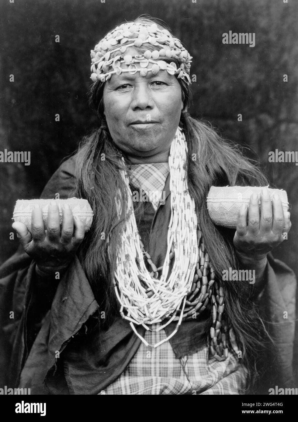 Hupa sciamano femminile, c1923. Athapascan Hupa donna della California nordoccidentale, ritratto a mezza lunghezza, in piedi, davanti, con archetti a conchiglia, collana e tiene in mano due cesti. Foto Stock