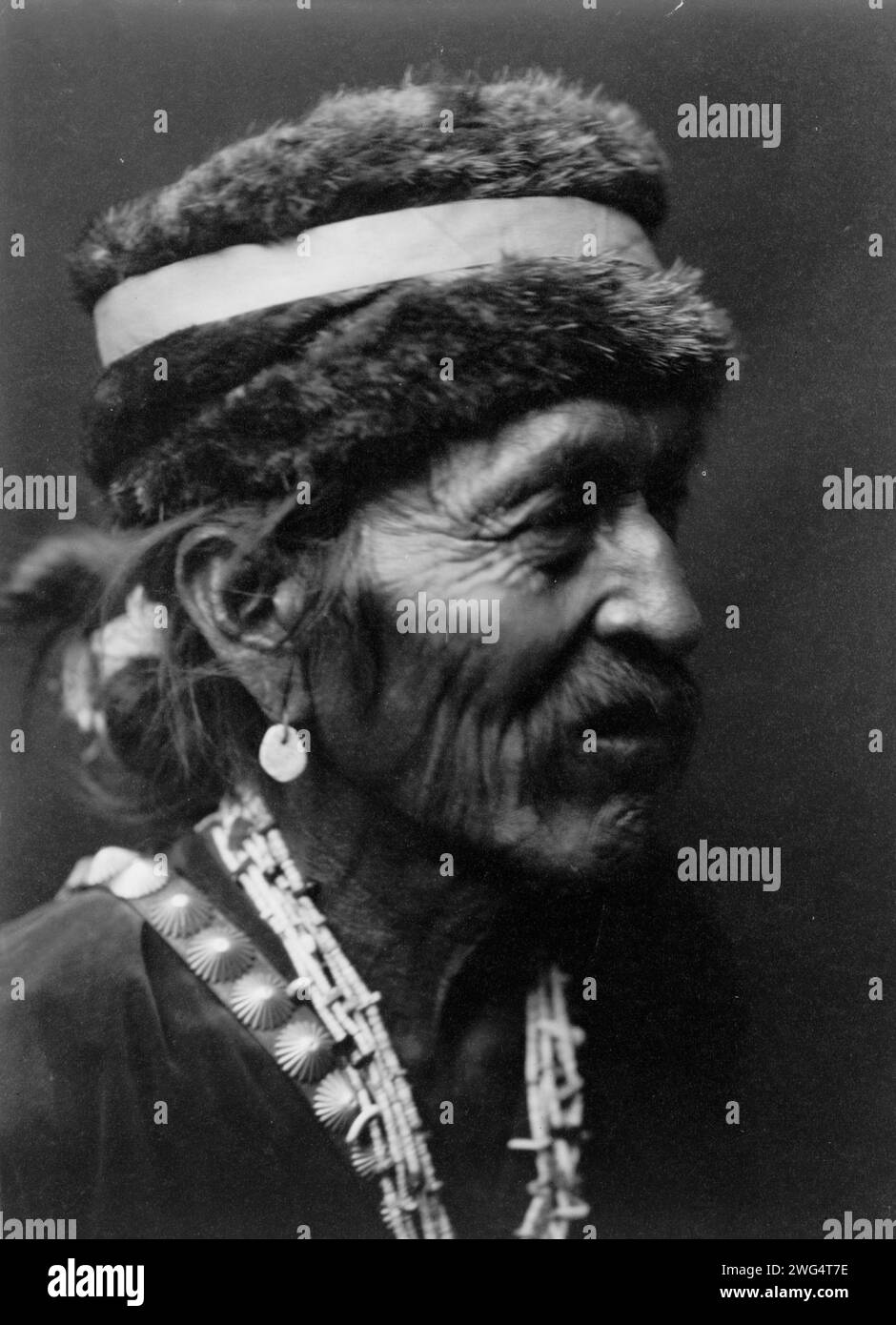 Navajo con berretto in pelliccia, c1905. Ritratto dell'uomo Navajo con la testa e le spalle rivolto verso destra, con berretto in pelliccia, orecchino turchese, collana con conchiglie, tracolla con bottoni in argento e camicia in velluto. Foto Stock
