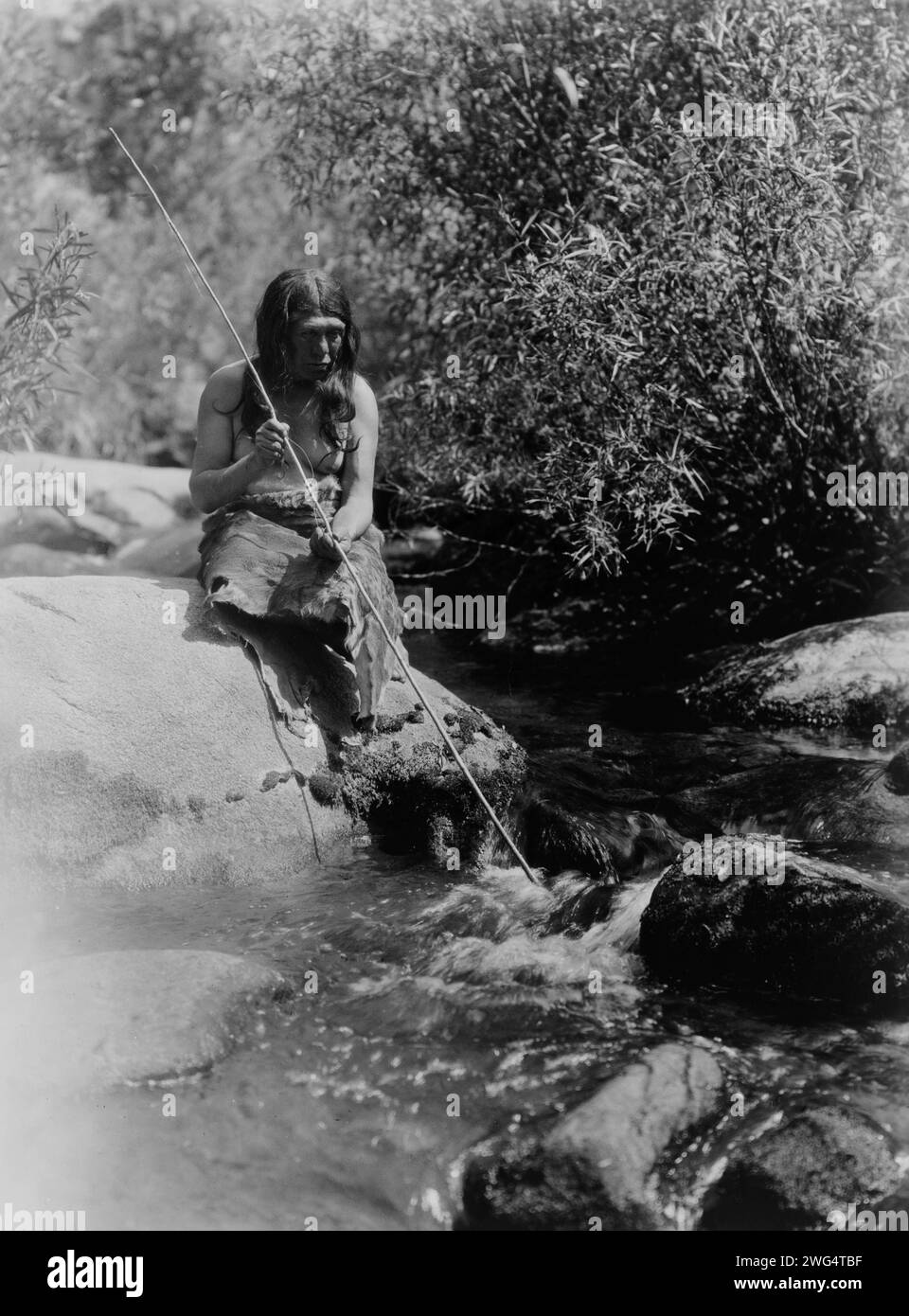 Sulla Merced-Southern Miwok, c1924. Un uomo Miwok che tiene la lancia, seduto su un masso in un torrente. Foto Stock