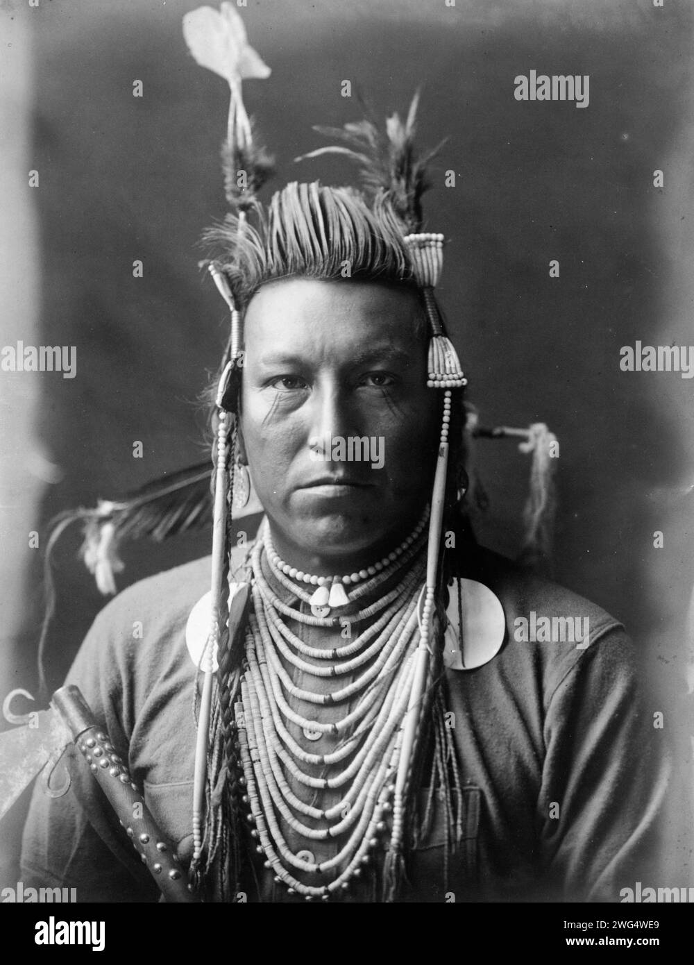 Swallow Bird, Crow Indian, Montana, ritratto testa e spalle, fronte, pompadour dipinto, copricapo di Apsaroke, camicia in flanella, perline sul petto, tubo con borchie tenuto davanti al braccio destro, c1908. Foto Stock