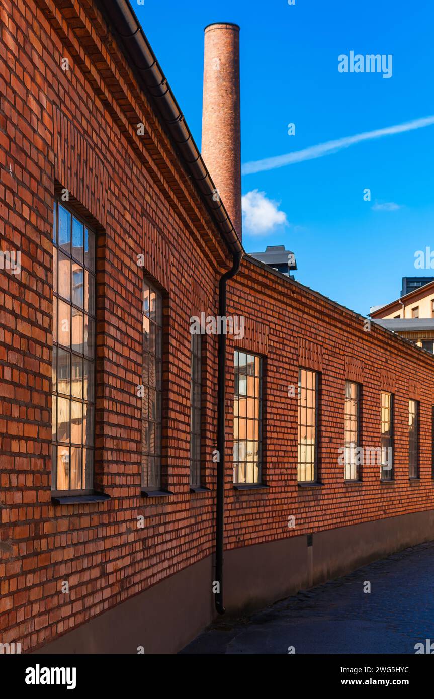 Un edificio industriale in mattoni rossi ben conservato, caratterizzato da un camino torreggiante contro un cielo blu brillante, evoca un senso di significato storico. Foto Stock