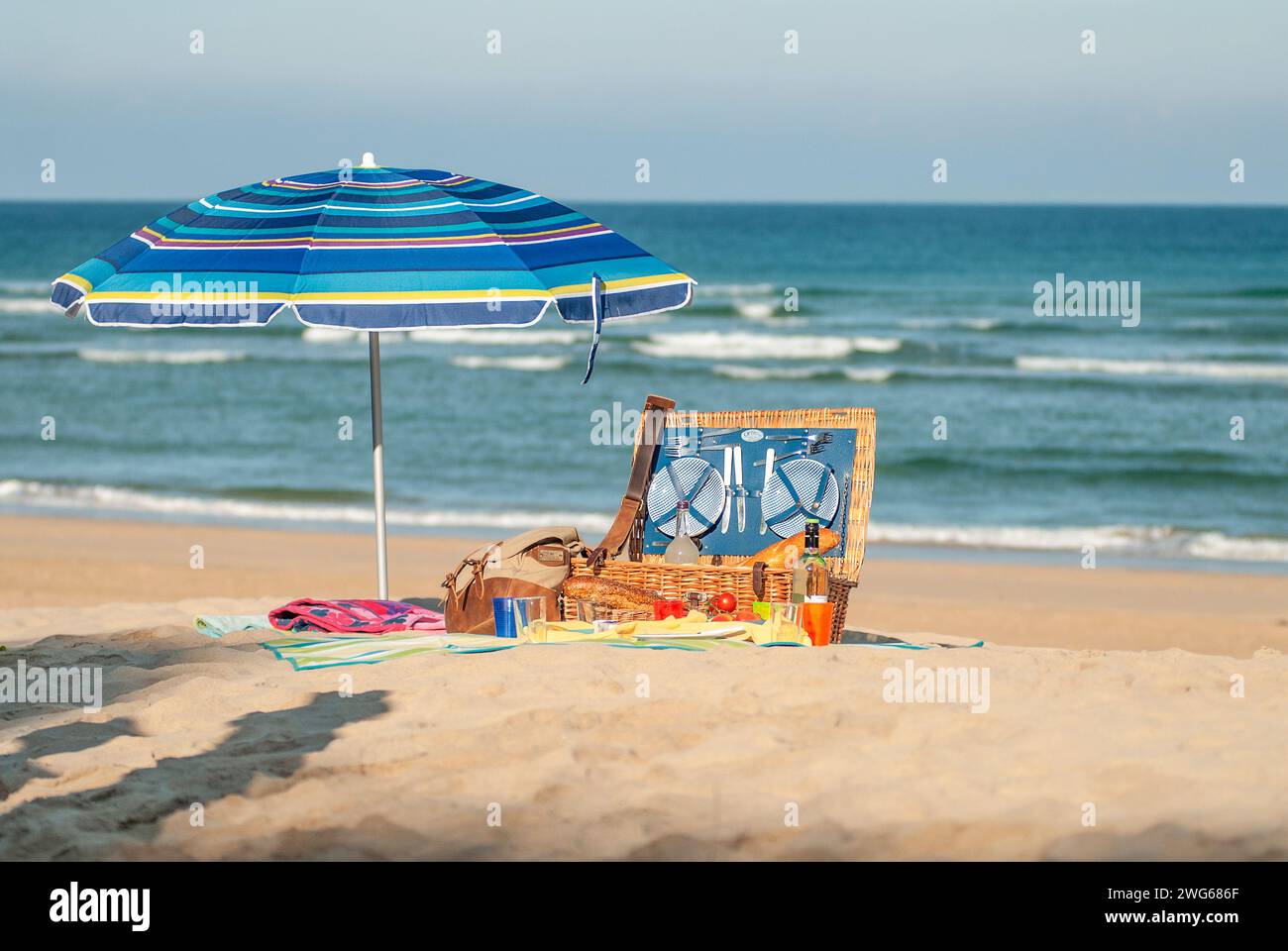 Un picnic disposto su una spiaggia vuota con cesto di vimini, tappeto e un ombrellone colorato. Foto Stock