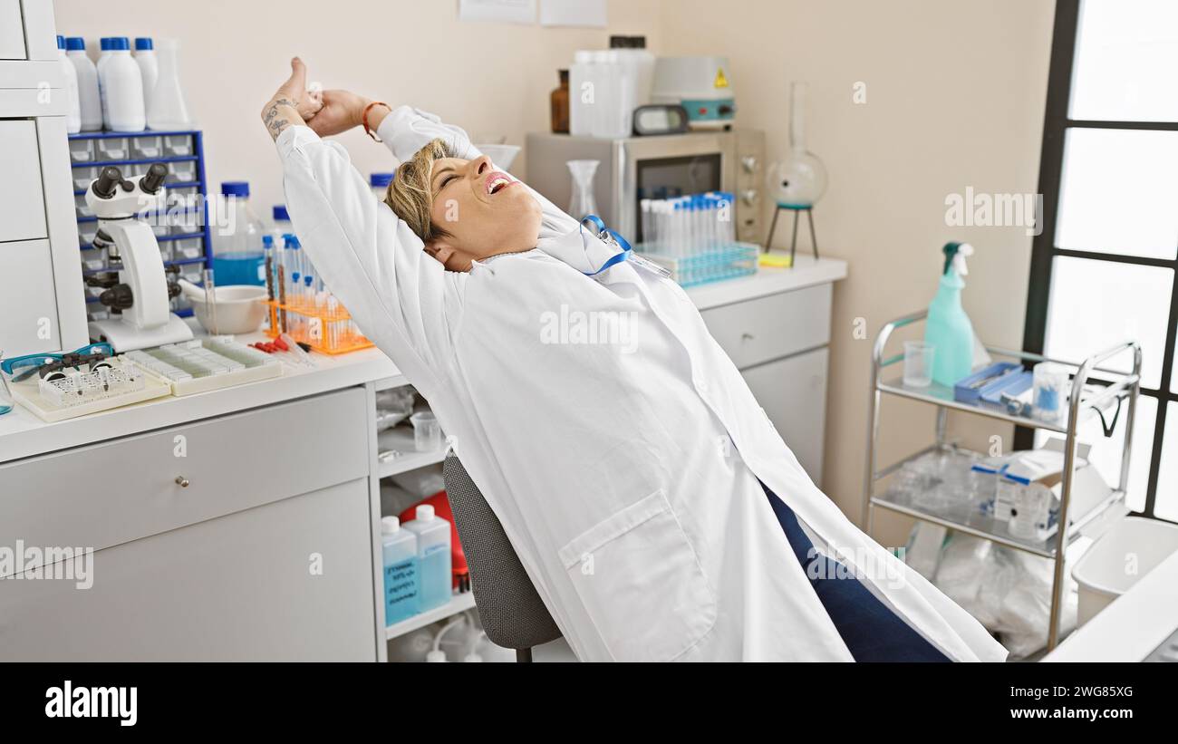 Donna ispanica rilassata con capelli biondi corti che si allungano in un camice bianco all'interno di un laboratorio. Foto Stock