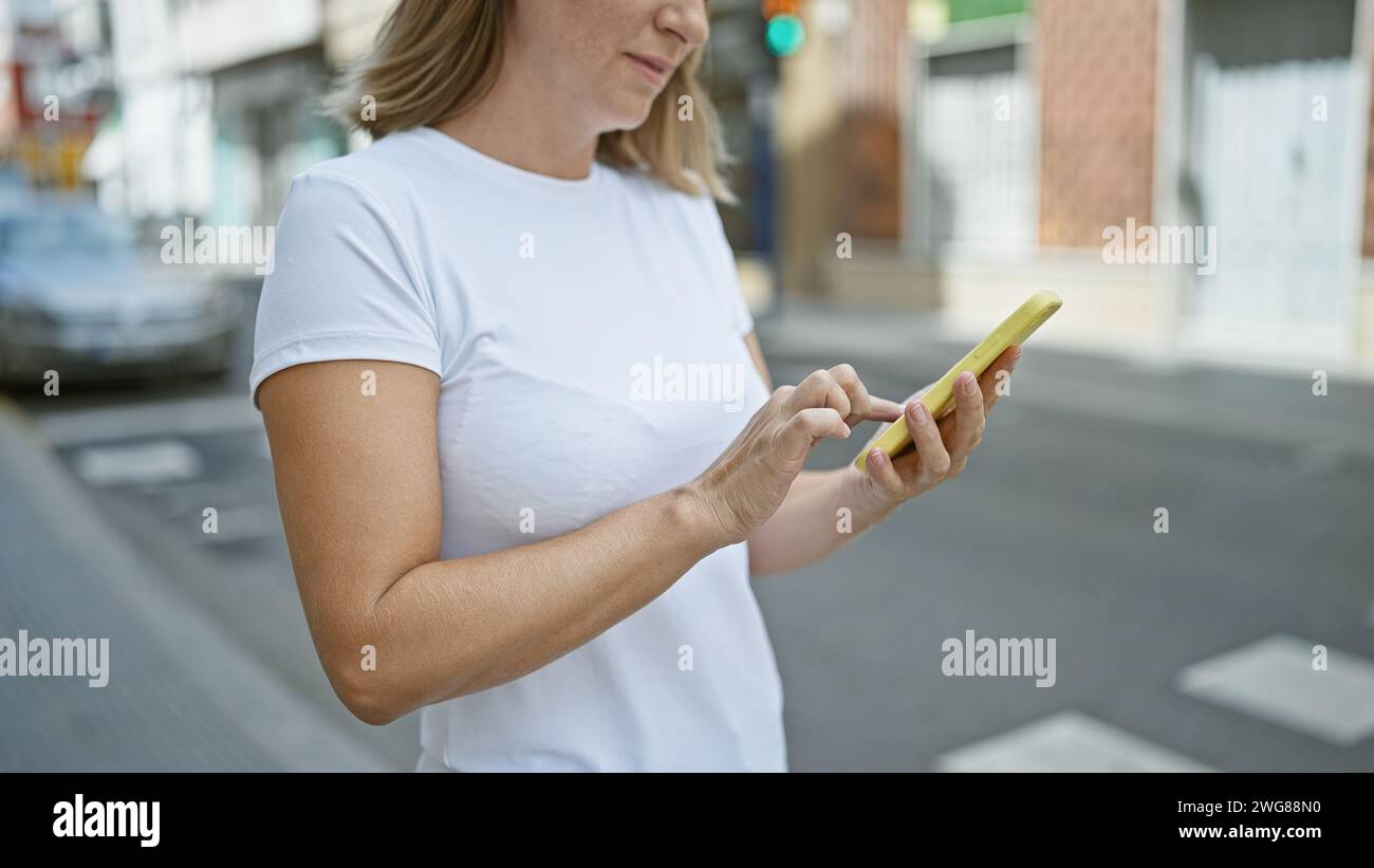 Elegante, bella e giovane donna bionda, si è concentrata seriamente sul inviare messaggi online sul suo smartphone, in piedi sulla soleggiata strada della città all'aperto, Livin Foto Stock