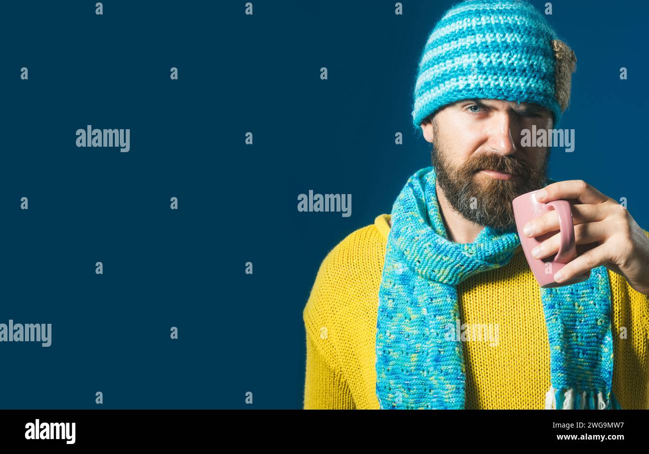 Uomo serio ed elegante con maglione a maglia e cappello avvolto in sciarpa con tazza di caffè o tè nel caffè. Bell'uomo barbuto in abiti invernali alla moda Foto Stock