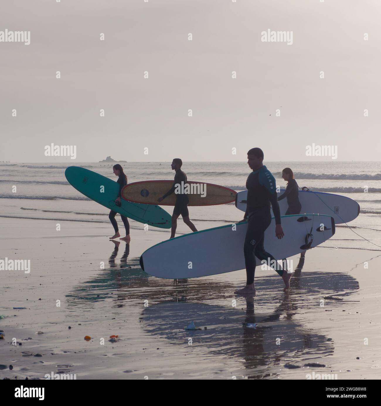 Un gruppo di surfisti lascia l'acqua trasportando le tavole che si riflettono sulla sabbia bagnata al tramonto a Essaouira, Marocco, 3 febbraio 2024 Foto Stock