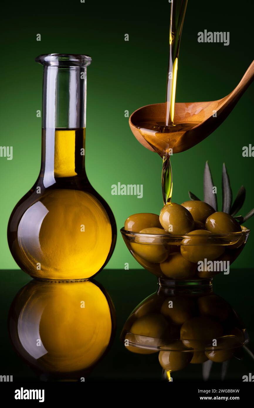dal cucchiaio di legno, in primo piano, l'olio extra vergine d'oliva viene versato in una ciotola con le olive. A parte una crosta di vetro con olio d'oliva Foto Stock
