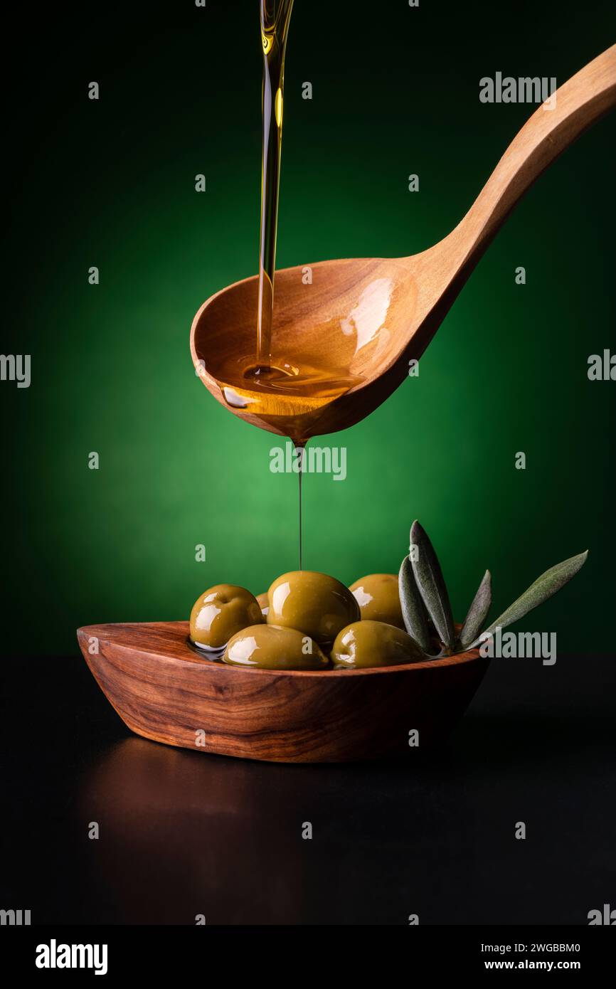 dal cucchiaio di legno, in primo piano, l'olio extra vergine d'oliva viene versato in una ciotola di legno d'oliva con olive Foto Stock