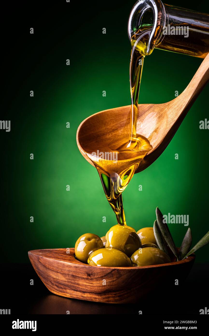 dal cucchiaio di legno, in primo piano, l'olio extra vergine d'oliva viene versato in una ciotola di legno d'oliva con olive Foto Stock