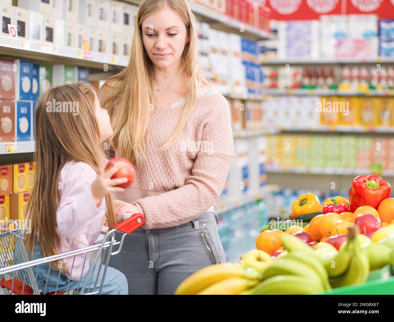 Felice e carina seduta su un carrello della spesa a comprare frutta e verdura fresca con sua madre al supermercato Foto Stock
