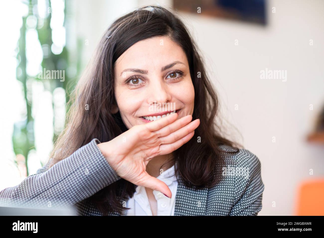 Donna d'affari sorridente seduta in un ufficio con la mano davanti al viso Foto Stock