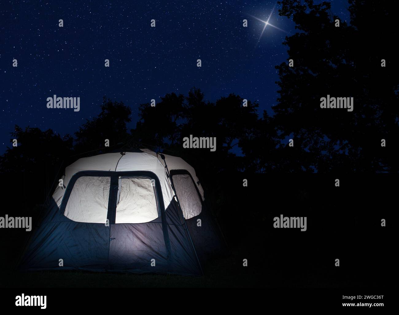 Grande tenda illuminata dall'interno, nei pressi di una foresta caratterizzata da stelle luminose, con una stella che risplende più luminosa nel cielo. Foto Stock