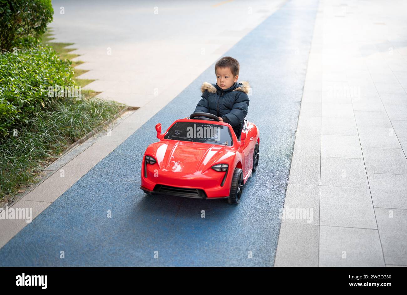 Un giovane bimbo multirazziale naviga con gioia sulla pista da corsa del quartiere a bordo di un'auto giocattolo sportiva rossa, sorridendo e assumendo il controllo di Foto Stock
